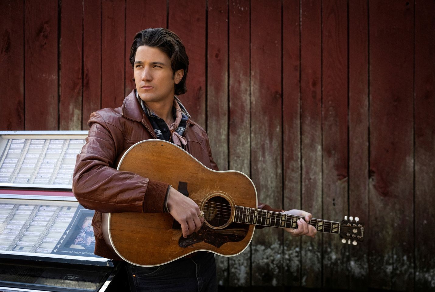 A man wearing a brown leather jacket holding an acoustic guitar